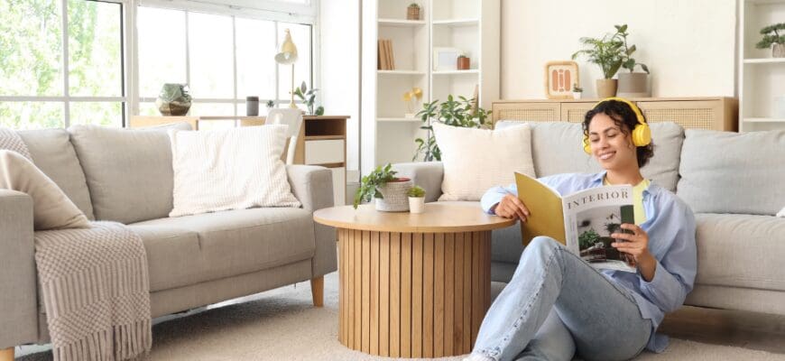 A woman sitting in a bright living room, wearing headphones and reading a magazine